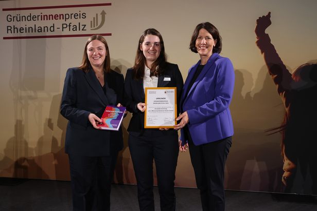 Drei Personen stehen vor einem Banner mit der Aufschrift „Gründerinnenpreis Rheinland-Pfalz“. Eine Person hält ein Buch mit dem Titel „INNOVATIONEN – Zukunft Jetzt“, eine andere eine gerahmte Urkunde. Die dritte Person steht daneben, alle befinden sich auf einer Bühne.