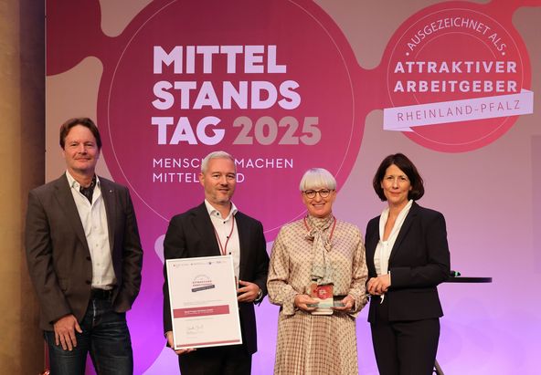 Vier Personen stehen auf einer B&uuml;hne vor einem Hintergrund mit den Aufschriften &bdquo;Mittelstandstag 2025&ldquo; und &bdquo;Attraktiver Arbeitgeber Rheinland‑Pfalz&ldquo;. Eine Person h&auml;lt eine gro&szlig;formatige Urkunde, eine weitere eine Auszeichnung. Rechts steht ein leerer Stehtisch.