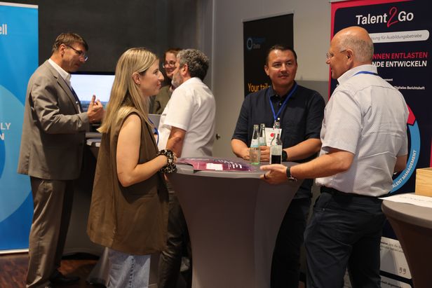 Sechs Personen im Gespräch bei Ausstellermarkt mit Stehtischen, Getränken und Roll-Ups von ‚Talent 2 Go‘ und ‚Oracle‘.