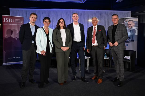 Sechs Personen stehen auf einer Bühne vor einem Hintergrund mit vertikalen Linien. Links ist ein Banner der Investitions- und Strukturbank Rheinland-Pfalz (ISB) mit dem Slogan ‚Zukunft. Fördern. Werte schaffen.‘ sichtbar