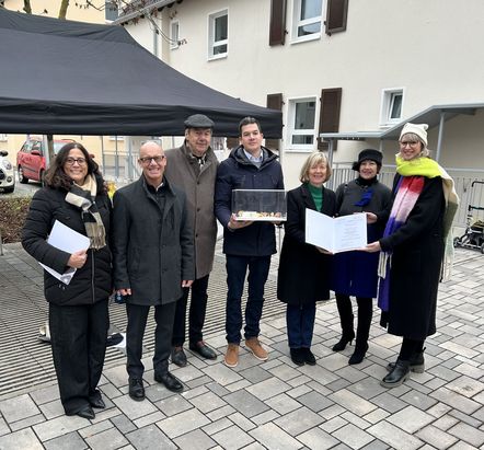 Sieben Personen stehen im Freien auf einem gepflasterten Platz vor einem hellen Geb&auml;ude mit braunen Fensterl&auml;den. Im Hintergrund ist ein schwarzes Zelt zu sehen. Zwei Personen in der vorderen Reihe halten ge&ouml;ffnete Mappen mit Dokumenten, eine weitere Person in der mittleren Reihe h&auml;lt ein rechteckiges Objekt. Alle tragen winterliche Kleidung.