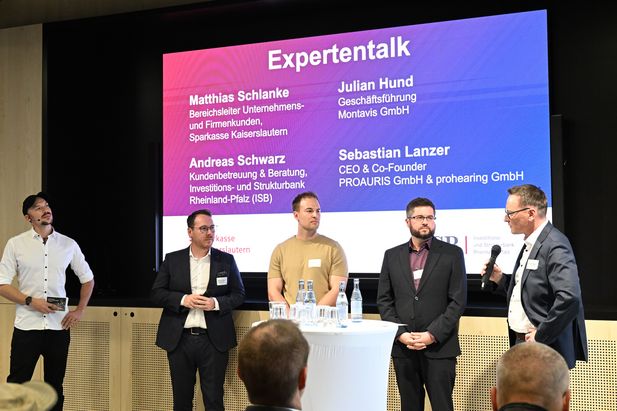 F&uuml;nf Personen stehen bei einer Expertenrunde auf einer B&uuml;hne vor Publikum. Im Hintergrund zeigt eine gro&szlig;e Pr&auml;sentationsfolie die Teilnehmenden und ihre Unternehmen.