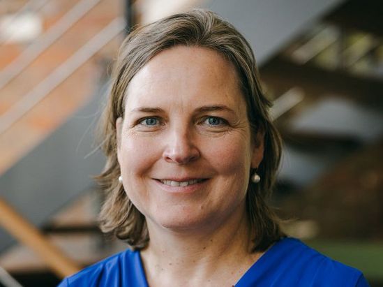 Eine Person steht vor einer modernen Innentreppe und tr&auml;gt ein leuchtend blaues Oberteil. Der Hintergrund zeigt unscharfe Holz‑ und Metallelemente einer offenen Geb&auml;udestruktur.