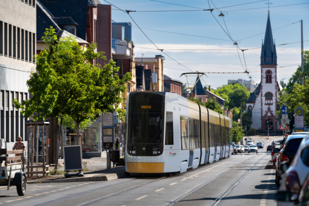 Moderne Stra&szlig;enbahn f&auml;hrt durch eine st&auml;dtische Stra&szlig;e in Mainz-Gonsenheim; im Hintergrund sind Wohn- und Gesch&auml;ftsgeb&auml;ude sowie eine Kirche zu sehen.