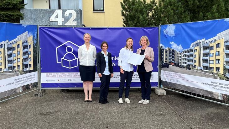 Vier Personen stehen vor einem großen lila Banner mit einem stilisierten Haus-Logo. Der Text auf dem Banner lautet: ‚Gefördert aus Mitteln des Bundes und des Landes Rheinland-Pfalz für die energetische Sanierung von Studentenwohnheimen‘. Links und rechts sind Bauzäune mit Bildern sanierter Wohngebäude zu sehen. Im Hintergrund ist die Zahl 42 auf einem grauen Stein erkennbar.