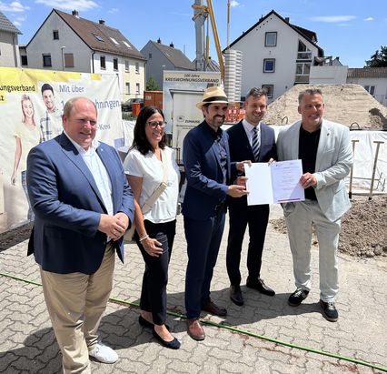 Eine Gruppe von sechs Personen steht im Freien auf einer gepflasterten Fläche vor einem Bauprojekt. Im Hintergrund sind Wohngebäude, ein Kran und ein Banner einer Wohnungsbau- oder Entwicklungsorganisation zu sehen. Eine Person hält ein Dokument, das im Mittelpunkt der Szene steht, was auf eine Übergabe oder ein offizielles Ereignis hinweist. Die Gruppe wirkt freundlich und posiert für ein Foto bei klarem Wetter. 