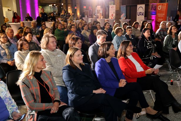 Ein Veranstaltungsraum mit vielen Personen, die in mehreren Reihen auf schwarzen Stühlen sitzen und nach vorne blicken. Im Hintergrund stehen Stehtische mit weißen Tischdecken sowie Banner und Roll-ups verschiedener Organisationen. Die Beleuchtung ist warm, mit violetten Lichtakzenten an den Wänden.