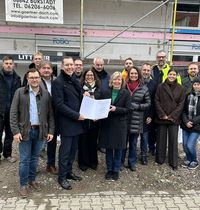 Vierzehn Personen stehen in zwei Reihen vor einem Gebäude mit Gerüst und Werbebanner. Im Vordergrund hält eine Person ein Dokument. Die Gruppe steht auf einem unbefestigten Untergrund neben einem Gehweg.