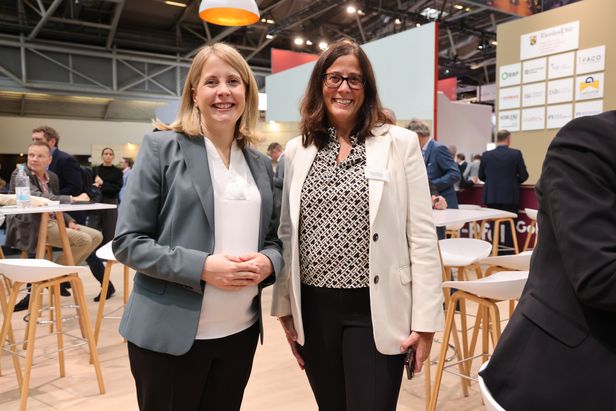 Zwei Personen in Businesskleidung stehen lächelnd in einem modernen, gut beleuchteten Veranstaltungsraum. Im Hintergrund sind weitere Teilnehmende an Stehtischen im Gespräch. Firmenlogos und Messestände sind sichtbar.