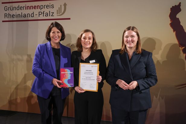 Drei Personen stehen vor einem Banner mit der Aufschrift „Gründerinnenpreis Rheinland-Pfalz“. Eine Person hält ein Buch mit dem Titel „INNOVATIONEN – Zukunft Jetzt“, eine andere eine gerahmte Urkunde. Die dritte Person steht daneben, alle befinden sich auf einer Bühne.
