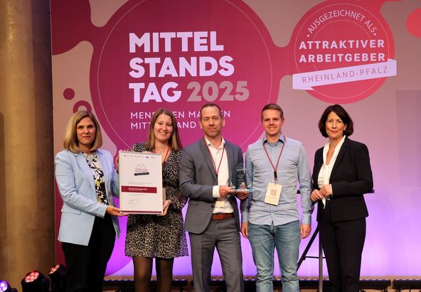 F&uuml;nf Personen stehen auf einer B&uuml;hne vor einem Hintergrund mit den Aufschriften &bdquo;Mittelstandstag 2025&ldquo; und &bdquo;Attraktiver Arbeitgeber Rheinland‑Pfalz&ldquo;. Eine Person h&auml;lt eine gro&szlig;formatige Urkunde, eine andere eine Auszeichnung. 