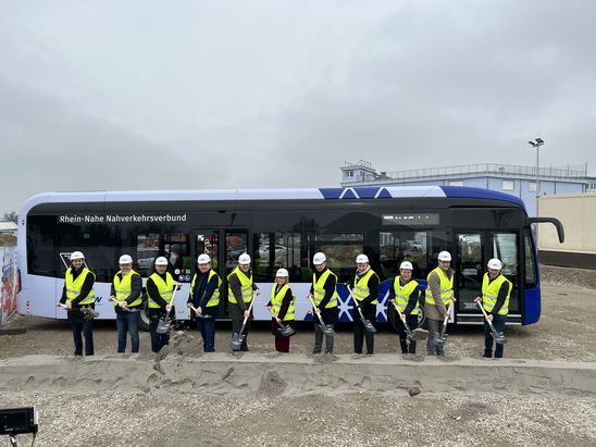 Gruppe von Personen mit Sicherheitswesten und Helmen steht mit Spaten vor einem Linienbus des Nahverkehrsverbunds auf einer Baustelle