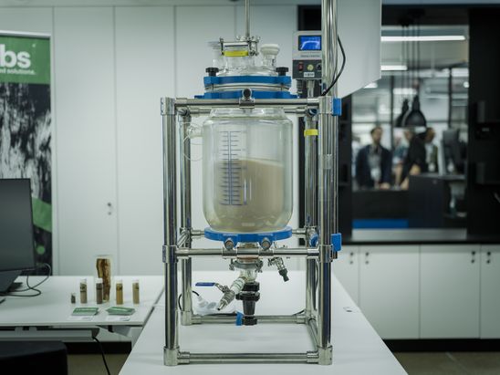 Gro&szlig;er transparenter Laborreaktor in einem Metallgestell, gef&uuml;llt mit heller Fl&uuml;ssigkeit, mit Schl&auml;uchen und Messger&auml;ten in einem Laborraum.