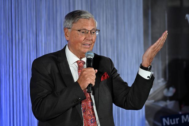 Person in Anzug spricht in ein Mikrofon und hebt eine Hand, vor einem Hintergrund mit vertikalen Linien