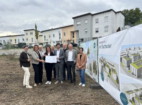 Gruppe von acht Personen auf einem Baugrundstück bei der Übergabe eines Förderbescheids vor einer Bautafel des Wohnbauprojekts „Am Bachacker“ in Heidesheim.