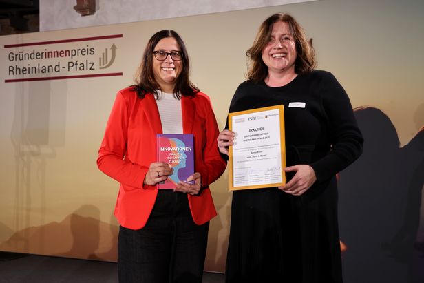 Zwei Personen stehen vor einem Banner mit der Aufschrift „Gründerinnenpreis Rheinland-Pfalz“. Eine Person hält ein Buch mit dem Titel „INNOVATIONEN – Zukunft Jetzt“, die andere eine gerahmte Urkunde. Beide stehen nebeneinander auf einer Bühne.