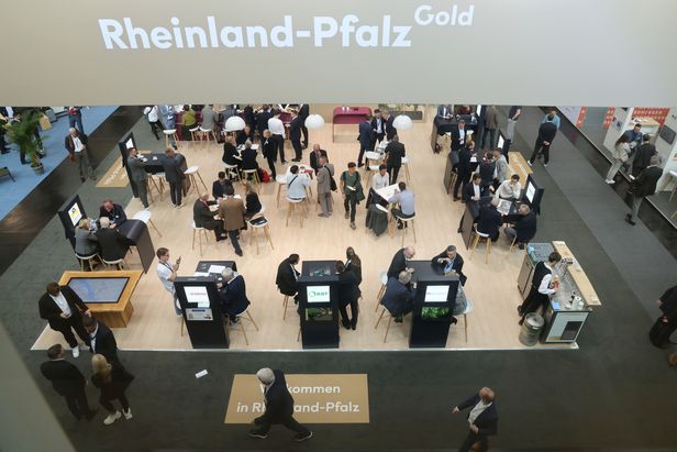 Blick von oben auf eine belebte Messehalle mit Gruppen von Menschen an Tischen. Ein großes Schild mit „Rheinland-Pfalz Gold“ hängt über dem Bereich. Am Boden steht „Willkommen in Rheinland-Pfalz“.