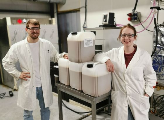 Zwei Personen in Laborkitteln stehen neben einem Tisch mit mehreren wei&szlig;en Kanistern in einer technischen Laborumgebung mit Rohren und Ger&auml;ten im Hintergrund.