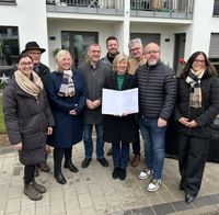 Acht Personen stehen auf einem gepflasterten Bereich vor einem weißen Gebäude mit Balkonen und Metallgeländern. Eine Person in der vorderen Reihe hält ein Dokument. Alle tragen winterliche Kleidung, einige haben Schals und Mäntel an.