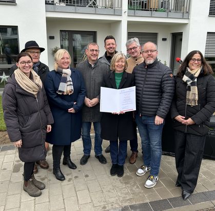 Acht Personen stehen auf einem gepflasterten Bereich vor einem wei&szlig;en Geb&auml;ude mit Balkonen und Metallgel&auml;ndern. Eine Person in der vorderen Reihe h&auml;lt ein Dokument. Alle tragen winterliche Kleidung, einige haben Schals und M&auml;ntel an.