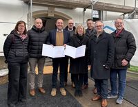 Förderbescheidübergabe an die Gemeinnützige Siedlungs-Gesellschaft (GSG) in Neuwied: Mehrere Personen stehen in einem Innenraum vor einem Banner. Eine Person überreicht einer anderen ein Dokument, während weitere Personen danebenstehen. Im Hintergrund sind ein Tisch und Informationsmaterial sichtbar.