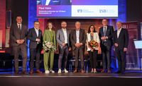 Acht Personen stehen auf einer Bühne bei einer Veranstaltung der ISB und der VR-Bank Südwestpfalz. Zwei Personen halten Blumensträuße, im Hintergrund sind die Logos beider Organisationen auf einer Präsentationsleinwand zu sehen.