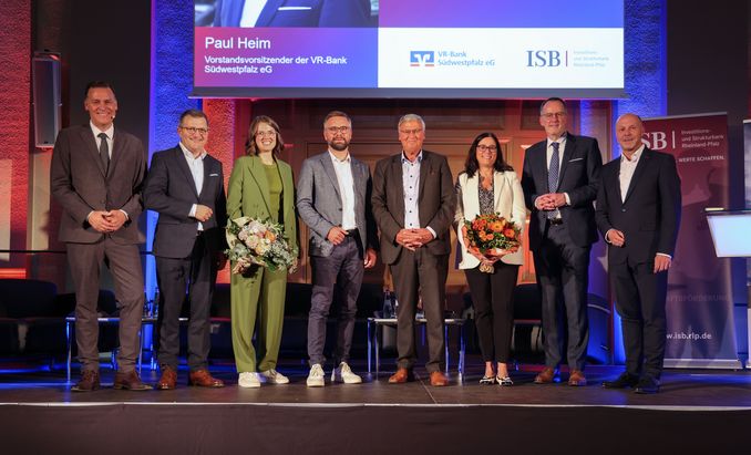 Acht Personen stehen auf einer B&uuml;hne bei einer Veranstaltung der ISB und der VR-Bank S&uuml;dwestpfalz. Zwei Personen halten Blumenstr&auml;u&szlig;e, im Hintergrund sind die Logos beider Organisationen auf einer Pr&auml;sentationsleinwand zu sehen.