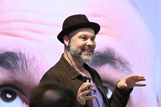 Person mit Hut und Headset-Mikrofon h&auml;lt einen Presenter in der Hand und spricht vor Publikum; im Hintergrund ist eine gro&szlig;formatige Projektion eines Gesichts zu sehen.