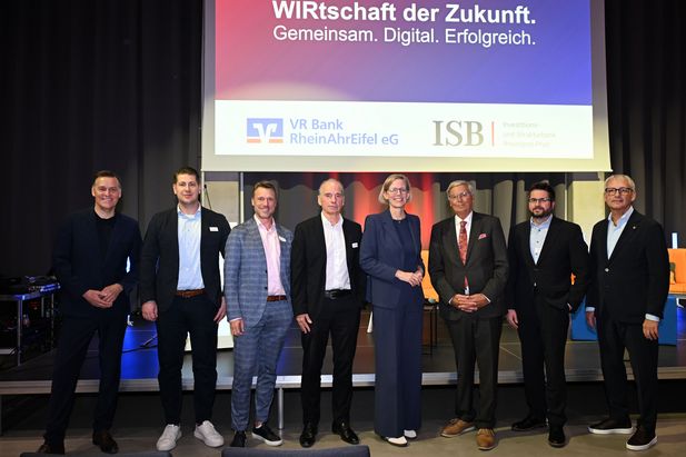 Acht Personen stehen auf einer Bühne vor einer großen Projektionsfläche. Auf der Leinwand sind die Logos der VR Bank RheinAhrEifel eG und der Investitions- und Strukturbank Rheinland-Pfalz (ISB) sowie der Schriftzug „WIRtschaft der Zukunft. Gemeinsam. Digital. Erfolgreich.“ zu sehen. Die Personen sind in Geschäfts- und Businesskleidung gekleidet und posieren für ein Gruppenfoto im Rahmen einer Veranstaltung.
