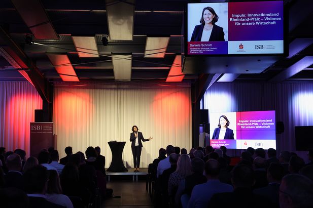 Eine Person steht auf einer Bühne vor einem weißen Vorhang und spricht zu einem Publikum. Über dem Publikum hängt ein Bildschirm mit der Aufschrift „Impuls: Innovationsland Rheinland-Pfalz – Visionen für unsere Wirtschaft“ sowie dem Namen „Daniela Schmitt“. Rechts neben der Bühne ist ein weiterer Bildschirm mit derselben Aufschrift und Logos der ISB und Sparkasse zu sehen.
