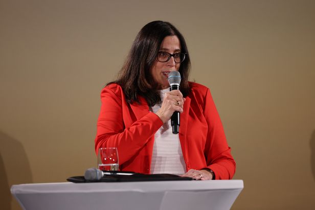 Eine Person steht hinter einem weißen Stehtisch und spricht in ein Handmikrofon. Auf dem Tisch steht ein Wasserglas. Der Hintergrund ist einfarbig beige.