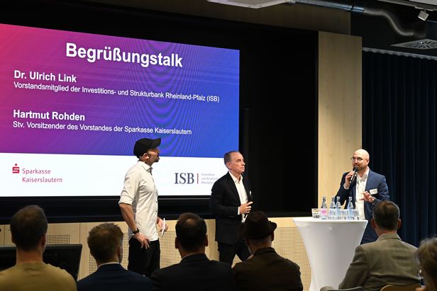 Drei Personen stehen bei einem Begrüßungsgespräch auf einer Bühne vor Publikum. Im Hintergrund ist eine Präsentationsfolie mit dem Titel „Begrüßungstalk“ zu sehen.