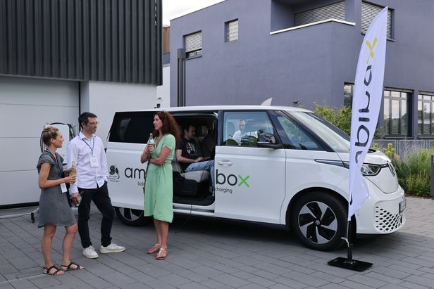 Vier Personen vor einem weißen Elektrofahrzeug mit der Aufschrift ‚ambibox‘; eine Person sitzt im Fahrzeug, drei stehen im Gespräch daneben. Rechts steht eine Fahne mit dem Schriftzug ‚ambibox‘, im Hintergrund moderne Gebäude